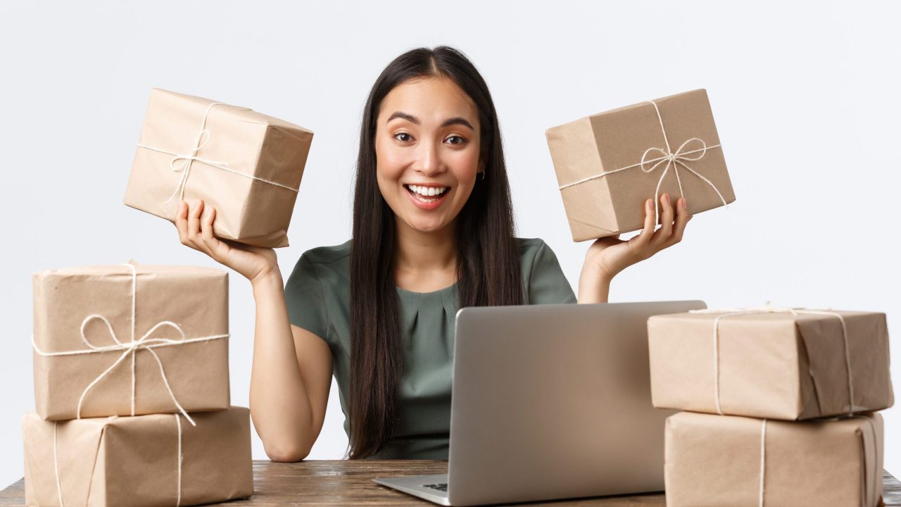 Small business owners, startup and e-commerce concept. Excited asian female entrepreneur sitting at desk with customer packages ready to delivery, smiling, answering client emails via laptop computer